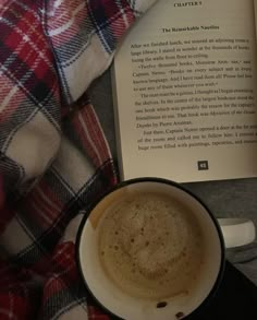 a cup of coffee sitting on top of a table next to an open book and blanket