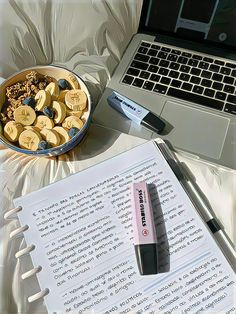 an open book and pen next to a bowl of bananas on a table with a laptop