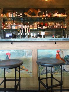 two stools in front of a bar with bottles on the shelves and paintings behind them