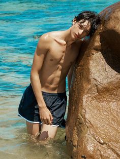 a young man standing in the water next to a large rock and wearing swim trunks