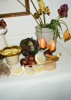 an assortment of fruits and vegetables sitting on a white table cloth next to a vase with flowers