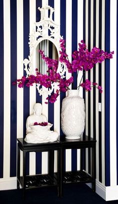 a white vase with purple flowers sitting on a table next to a mirror and striped wall