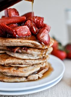 a stack of pancakes with syrup being poured on top and strawberries in the background