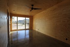 an empty room with a ceiling fan and large window overlooking the desert outside, in front of a wooden wall that is partially covered by plywood paneling