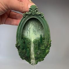 a hand holding up a small piece of art with trees and water coming out of it