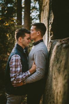 two men standing next to each other in front of some trees and one is kissing the other