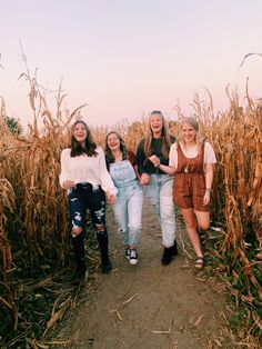 four girls walking through a corn field holding hands and smiling at the camera while wearing overalls