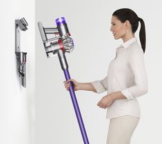 a woman holding a purple and silver vacuum in her right hand while standing next to a white wall