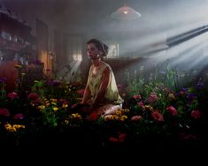 a woman sitting on the ground surrounded by flowers in front of a light that is shining down