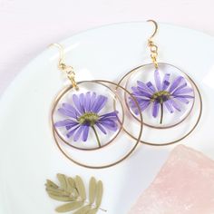 two purple flowers are hanging from hoop earrings on a white plate next to some leaves