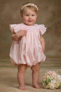 a baby girl in a pink dress standing next to a flower bouquet and smiling at the camera