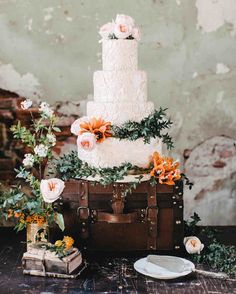 a white wedding cake with flowers and greenery on top sits on a table in front of an old brick wall