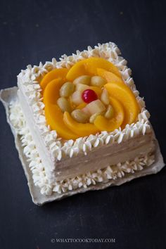 a square cake with white frosting and fruit toppings on the top, sitting on a black surface