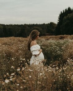 a pregnant woman standing in a field with wildflowers and trees behind her, wearing a white dress