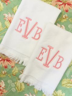 two towels with embroidered initials on them sitting on a floral print table cloth, one is white and the other has pink letters