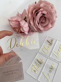 someone is holding up some business cards with gold lettering on them and pink flowers in the background