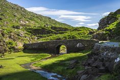 an old stone bridge over a stream in the middle of a green valley surrounded by mountains