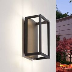 an outdoor light mounted on the side of a white wall next to a tree with pink flowers