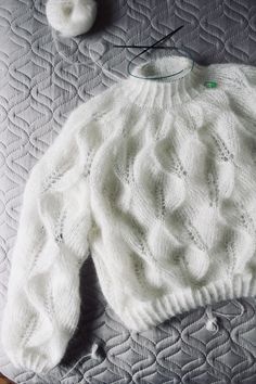 a white knitted sweater sitting on top of a bed next to a ball of yarn