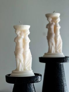 two white candles sitting on top of each other in front of a gray wall and black stand