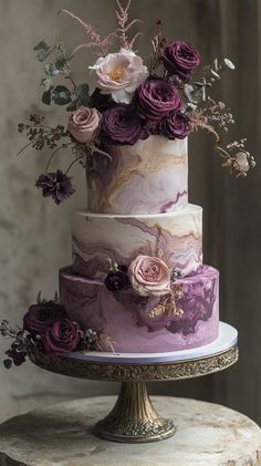 a three tiered cake with purple flowers and greenery on the top is sitting on a pedestal