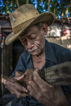 an old man wearing a hat and holding a piece of wood in his hands while sitting down