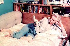 a woman laying in bed reading a book with bookshelves and pictures on the wall behind her