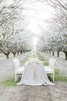 an outdoor table with white chairs and flowers on it in the middle of some trees