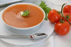 a white plate topped with a bowl of tomato soup next to two tomatoes and a spoon