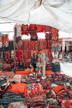 two people and a child standing in front of a pile of cloths at an outdoor market