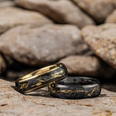 two black and gold wedding bands sitting on top of each other in front of some rocks