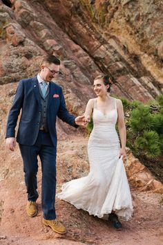 a bride and groom holding hands while walking on a trail in the mountains near some rocks