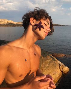 a shirtless man sitting by the water with a starfish on his forehead