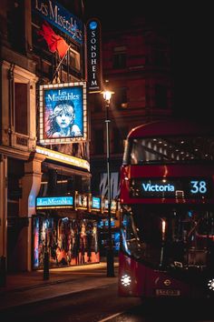 a red double decker bus driving down a street at night with people walking on the sidewalk