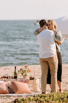 a man and woman embracing on the beach with an umbrella over their head, next to a picnic table