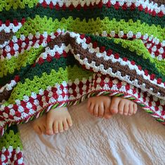 a baby laying under a crocheted blanket on top of a white bed covered in green, red and brown blankets