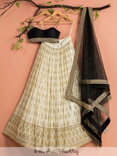 A three-piece ivory and black shimmer lehenga set from the Anisha Shetty collection. This gorgeous ivory georgette lehenga is paired with a raw silk black bustier blouse. The ivory lehenga is enhanced with heavy sequence work all over with scalloped edging. The black blouse has a beautiful sequence-zari work & a sequin tassel tie-up at the back. The outfit is completed with a net black dupatta with heavy sequence and badla work. Cheap Fitted Lehenga For Eid, Lehenga With Black Blouse, Black Shimmer Lehenga, Shimmer Lehenga, Badla Work, Ivory Lehenga, Bustier Blouse, Sequence Lehenga, Simple Lehenga