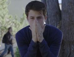 a young man covers his face with his hands as he walks past trees and people