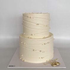 a three tiered white cake with pearls on the side and two gold wedding rings next to it