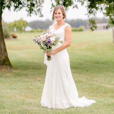 a woman in a white dress holding a bouquet of flowers standing on the grass near a tree