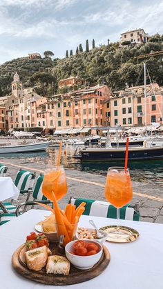 two glasses of wine and some food on a table near the water with boats in the background