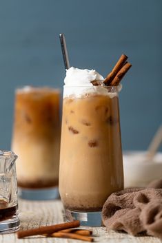 two glasses filled with drinks sitting on top of a table next to cinnamon sticks and whipped cream