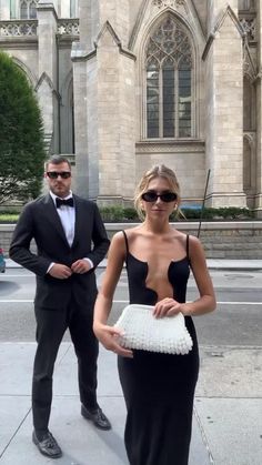 a woman in a black dress and a man in a tuxedo stand on the sidewalk