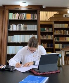 a woman sitting at a table in front of a laptop computer and writing on a piece of paper