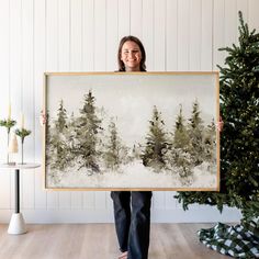 a woman holding up a painting in front of a christmas tree with snow on it