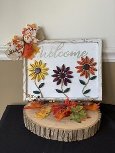 a welcome sign with flowers and leaves on top of a tree stump in front of a wall