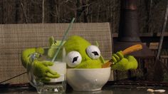 a green stuffed animal holding a spoon in a bowl next to a glass of milk