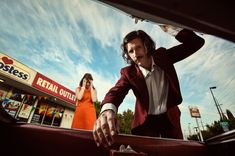 a man in a red suit and white shirt standing next to a woman with her hand on the hood of a car