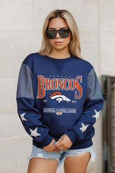 a woman wearing a blue denver football sweatshirt and denim cut - off shorts is standing in front of a white brick wall