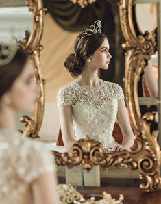 a woman in a white dress looking at her reflection in a gold framed mirror while wearing a tiara